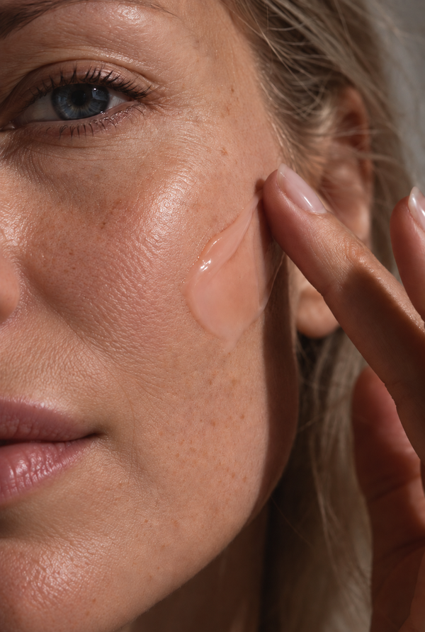 Close-up of a woman's face with a translucent gel or cream swatch applied to her cheek, with her finger touching the product area.